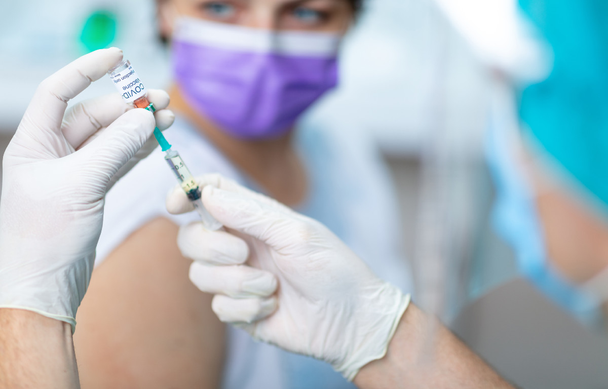 Foreground of needle with Covid vaccine being held; background, woman with sleeve rolled up awaiting shot. 