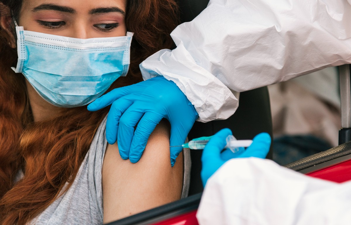 Young woman wearing mask looks down as vaccine injected into her arm
