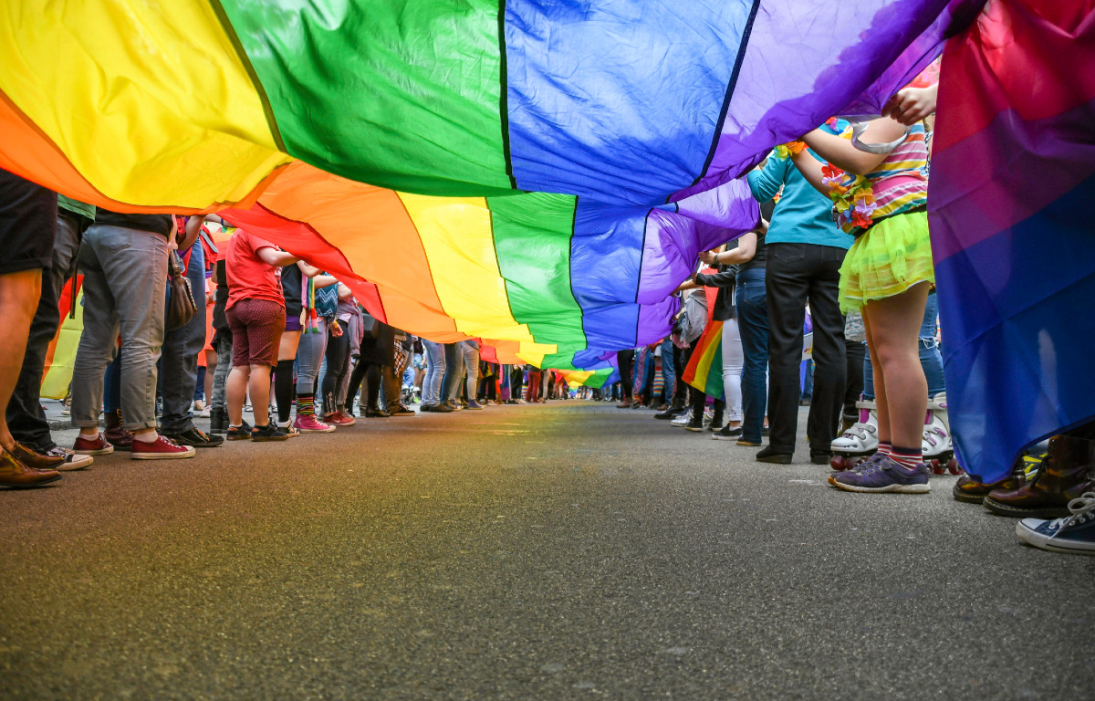 Pride flag lengthwise being held by numerous people facing away from camera on both sides