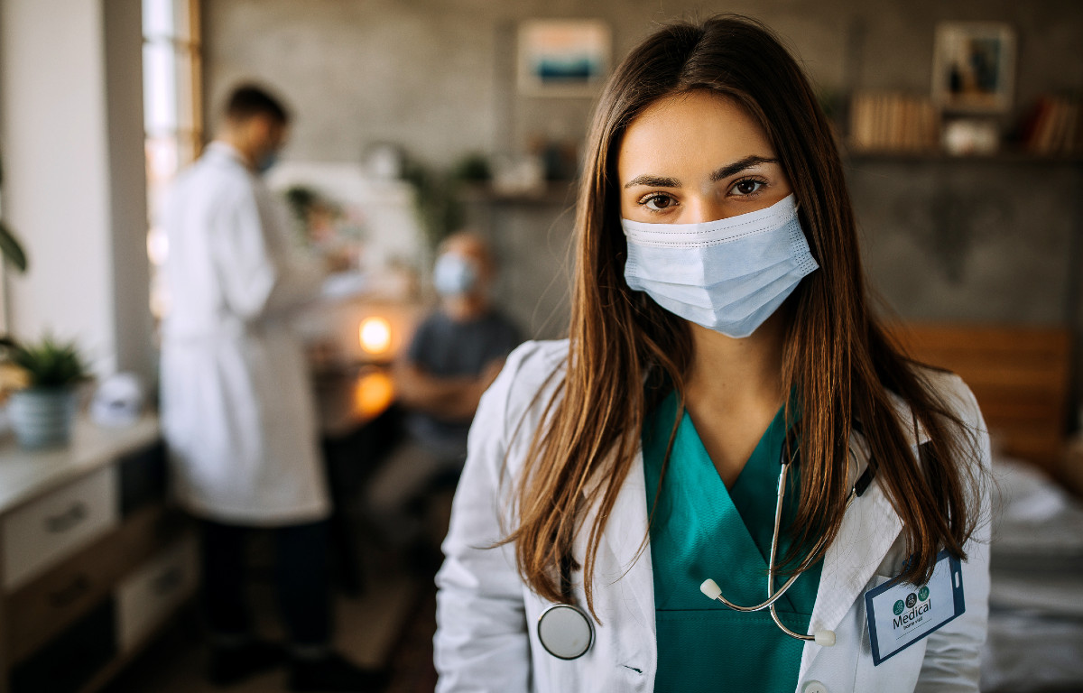 Young woman doctor wearing mask looking at camera with other doctors (blurry) talking in background